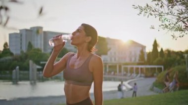 Yorgun bir bayan akşam sporundan sonra su dengesini tazeliyor. Rahat formda bir kadın güneş batımı parkında spor şişesinden sıvı içer. Mutlu sporcu kadın moladan zevk alıyor. 