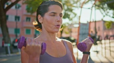 Ciddi sporcu kadın güneş ışınları caddesindeki spor salonunda dambılları kaldırıyor. Mutlu gülümseyen kadın, yalnız başına rüzgarlı yaz şehrinde kaslarını çalıştıran kardiyo antrenmanından zevk alıyor. Spor etkinliği kavramını biler 