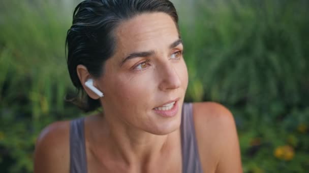 Yogi女士在夏季公园的特写镜头边讲耳机 在绿草背景下使用现代无线耳塞远程说话的成熟女士保持冷静 瑜伽教练在网上教授健身课 — 图库视频影像