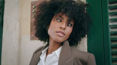 Gorgeous young model posing leaning on building wall close up. Beautiful african american woman with lush curly hairstyle standing outdoors looking camera. Attractive exotic girl relaxing on street.