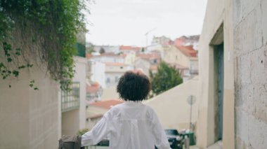 Playful happy woman running down city street holding jacket. Carefree african american girl rejoicing beautiful weekend in town. Smiling attractive lady tourist enjoying summer walk relaxing outdoors.