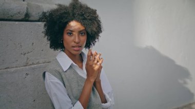 Gentle attractive girl standing enjoy sunlight near stone wall close up. Attractive curly african american woman posing outdoors wearing white shirt. Stylish beautiful brunette relax looking camera.