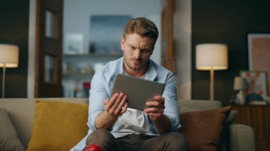 Odaklanmış hipster muayene sehpası bilgisayar oturma koltuğu. Ciddi bir adam oturma odasının içinde dijital cihaz kullanarak yeni bir uygulama deniyor. Akşam apartmanında zoom yapan adam merak ediyor. 