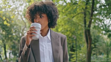 Relaxed stylish woman walking city park at work break enjoying coffee takeaway close up. Confident african american business lady drinking beverage in paper cup. Attractive girl looking smartphone.