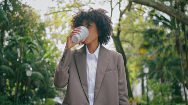 Young stylish business lady going at city park alley with paper cup hot coffee closeup. Confident african american woman holding drink takeaway walking alone. Curly girl in elegant suit looking phone.