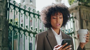 Attractive curly business lady talking phone standing at iron gates with coffee cup close up. Elegant african american woman wearing stylish suit using mobile outdoors. Confident girl holding drink.