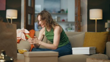 Kanepede akıllı telefon kullanan mutlu bir kadın. Kıyafetlerle dolu kanepeye duygusal olarak sevinen alışveriş meraklısı. Gülümseyen neşeli bayan alışverişlerin etrafında dans ederken eğleniyor. Başarılı alışveriş kavramı