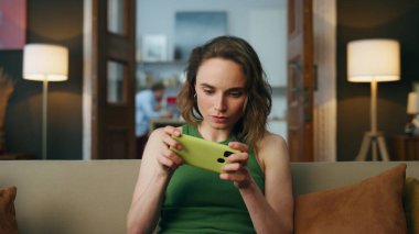 Evde cep telefonuyla oynayan odaklanmış bir oyuncu. Kaygısız kadın akıllı telefon oyunlarından zevk alıyor hafta sonu kanepede dinleniyor. Ev zoom 'unda kanepeye oturup eğlenen kadın dahil. Kız eğleniyor.