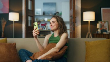 Dost canlısı, el sallayan, akıllı telefonlu bir bayan. Rahat bir kadın kanepede oturmuş, video odasında arkadaşını selamlıyor, zum yapıyor. Gülümseyen kız cep telefonu tutuyor. Kanepede tek başına konuşmanın tadını çıkarıyor.