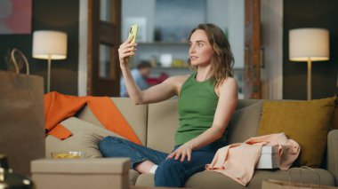 Gülümseyen blogcu evde yayın yapıyor. Çekici modern kadın telefon kullanarak alışveriş vlog 'u kaydediyor. Kanepede el kol hareketi yapan aktif bir bayan. Gülümseyen kız tek başına akıllı telefon içeriğini kaydediyor