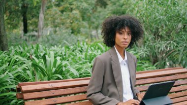 Attractive confident girl freelancer working in beautiful park close up. Stylish curly african american woman typing on laptop sitting bench. Young business lady using wireless computer outdoors.