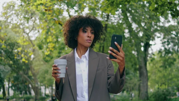 Confident stylish woman talking on mobile camera walking city park with paper cup coffee takeaway close up. African american girl speaking at video call outdoors. Business lady have online conference.