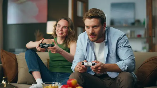 Two people competing arcade game at living room. Proud woman winner looking at loosing husband. Confused man feeling embarrassed sitting couch. Millennial couple playing videogames at home place