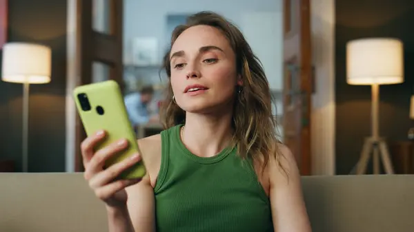 Dost canlısı bir kız cep telefonunu arıyor. Gülümseyen kadın karşılama eli ile konuşuyor cep telefonuyla görüntülü sohbet yakınlaştırması. Mutlu bayan oturma odasında sanal telefon görüşmesi yapıyor.