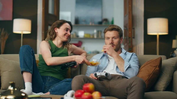 Smiling woman giving snacks to man at beige sofa interior. Angry loosing guy taking second try starting playing game. Purposeful gamer holding remote controller competing wife. Weekend entertainment
