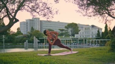 Atletik kadın Alacakaranlık Parkı 'nda yoga pozu veriyor. Sportif, olgun bir kadın, yeşil doğada paspasın üzerinde uzun bir yan açı pozisyonu alıyor. Yogi antrenörü dışarıda vücut germe egzersizi yapıyor. 