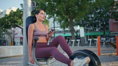 Spor kadın, parkta pedal çevirirken cep telefonunu izliyor. Orta yaşlı aktif sporcu kadın akıllı telefon mesajı atıyor açık hava spor salonunda tek başına egzersiz yapıyor. Sağlıklı yaşam tarzı kavramı