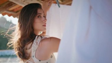 Rüzgarlı teras portresine çamaşır asan seksi bir kadın. Romantik bir kadın ev bahçesinde ıslak kıyafetlere dokunuyor. Güneşli yaz gününde beyaz çarşaf havası kuruyor. Ev içi rutin