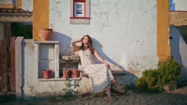 Nazik kız, güneş ışığını ısıtıyor, eski evin duvarına yaslanıyor. Romantik elbiseli mutlu bir kadın sıcak rüzgarın tadını çıkarıyor. Genç esmer kadın yaz güneşinde saç stiline dokunuyor. 