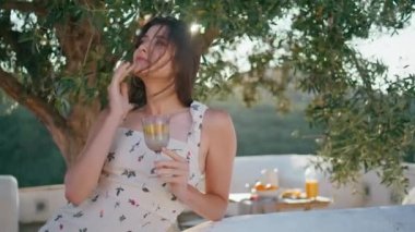 Rüzgarlı saçlı bayan terası gevşetiyor cam içki tutuyor. Romantik bir kadın yaz sabahı kameraya dokunuyor. Rahat model tek başına siestanın tadını çıkarıyor. Tatil yaşam tarzı kavramı
