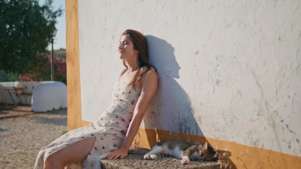 ベンチで寝ている猫の近くでシエスタを楽しむラジーモデル ハッピー笑顔の女性が日光浴して 地中海の古い町を一人でリラックス 夏に休むロマンチックなドレスのケアフリーガール — ストック動画