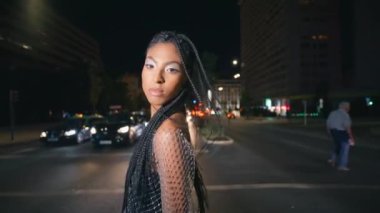 Modaya uygun esmer kız gece trafiğini geçiyor. Muhteşem stil sahibi bayan şehir fotoğraflarına bakan kameralara bakmaktan zevk alıyor. Moda kadını şehrin arka planında yalnız poz veriyor.