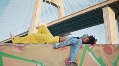 Şehir köprüsünün önünde uzanan siyah saçlı kız. Rahat Afro-Amerikan genç bayan duvarda graffitiyle rahatlıyor. Şehrin arka planında poz veren modaya uygun kıvırcık kadın model..