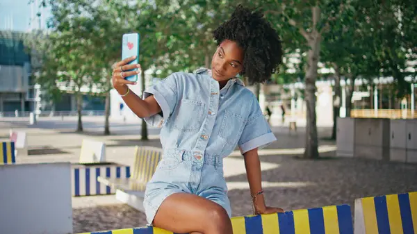 African american girl blogging using smartphone sitting sunny street. Smiling female blogger looking cellphone camera streaming online outdoors. Curly stylish teenager recording live video on phone.
