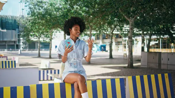 Teenager talking at video meeting by modern cellphone sitting colorful street bench. Happy african american girl smiling at online call by smartphone outdoors. Cute teen speaking to mobile camera.