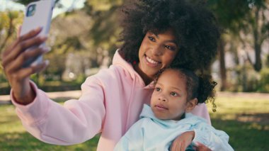 Annem Green Park 'ta sevimli küçük kızıyla selfie çekiyor. Mutlu Afro-Amerikan kadın kıvırcık çocukla çayırda otururken fotoğraf çekiyor. Akıllı telefon kamerasına gülümseyen neşeli kızlar.