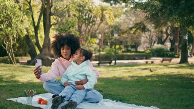 Gülümseyen anne, kızıyla birlikte modern akıllı telefonu kullanıyor. Şirin Afrikalı Amerikalı kız dışarıda telefon kamerasına bakarak mutlu anneyi öpüyor. Parkta neşeli aile blogları.