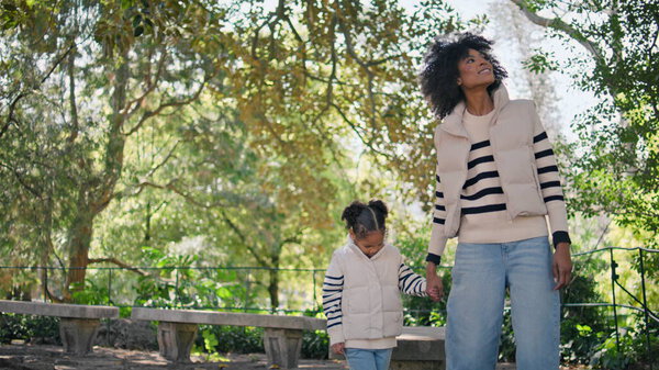 Mother leading little daughter by hand walking on beautiful green park. Smiling african american woman looking around enjoy family weekend. Cute pretty girl strolling holding mom hand. Leisure concept