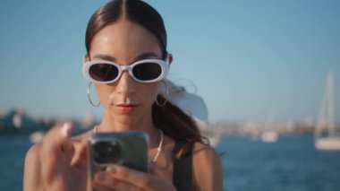 Sahildeki son moda bayan mesajlaşması. Güneş gözlüklerini çıkartıyor. Yakından. Güneşli deniz kıyısında gülümseyen güzel, mutlu bir kadın. Güzel kız cep telefonuyla sahilde mesaj okuyor..