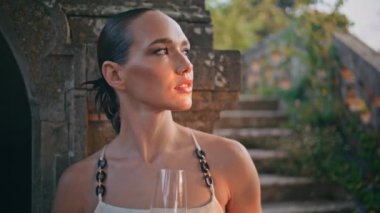 Lüks model, dışarıdaki eski taş sarayda akşamın tadını çıkar. Şarap kadehi tutan muhteşem genç bayan antik şato yaz akşamının dışında dinleniyor. Şarap kadehi görünümlü kameralı rahat bir kadın..