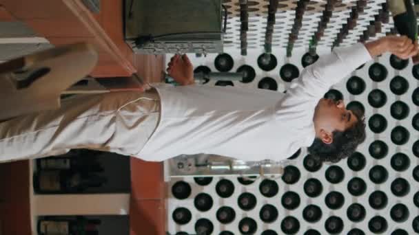 ワイナリーコレクションからワインボトルを取る若いカーリー男垂直方向 アルコール飲料のイタリアのソムリエ読書ラベル レストランでの夕食に美味しい飲み物を選ぶ自信あふれるグルメ — ストック動画