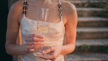 Tanımlanamayan bir kadın elinde şampanya bardağıyla akşam güneşinin altında dinleniyor. Lezzetli beyaz şarapla doldurulmuş zarif kadehli kız elleri. Şık bayan eski binanın dışında parti yapıyor..
