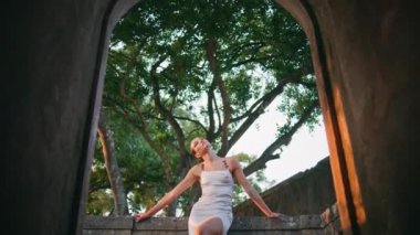 Kemerden taş çitlere yaslanmış antik bir binayı poz veren şık bir kadın. Terk edilmiş bir kalenin balkonunda duran muhteşem lüks kadın. Güzel, rahat bir kız bahçedeki ağaçların tadını çıkarıyor.