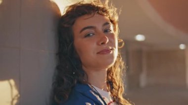 Akşam güneşinde dışarıda poz veren güzel bir melomanyağın portresi. Kulaklıkla müzik dinleyen kıvırcık modacı kadın şehir caddesinde duruyor. Mutlu kız şarkıları seviyor. Güneşli hafta sonlarında dinleniyor. Yakın plan..