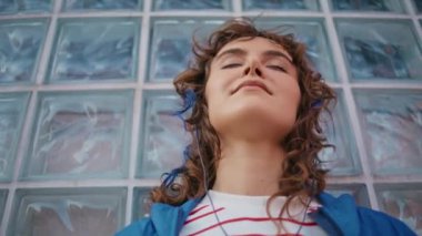Yakın çekim, cam duvar binasına dayanan kulaklıkla müzik dinleyen gevşek melodi manyağı. Kıvırcık sakin bir kızın portresi gözlerini kapatıyor ve güzel bir sesin tadını çıkarıyor. Sokakta kulaklıkla poz veren kaygısız kadın.