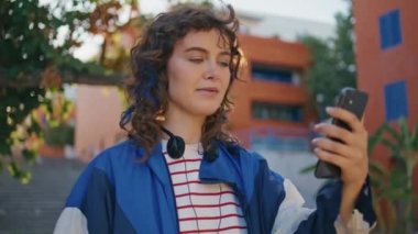 Şehir sokağındaki akıllı telefondan son moda kız videosu kapanıyor. Cep telefonuyla konuşan mutlu kıvırcık kadın şehir merkezinde yürüyor. Modern hipster 'ın dışarıda sanal bir sohbeti var..