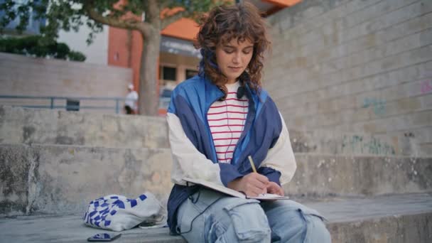 一个浪漫的女孩独自在市区附近写笔记本 坐在水泥楼梯上 在记事本上做笔记的卷曲放松的女人 多愁善感的年轻学生在户外做家庭作业简编 青少年生活方式 — 图库视频影像