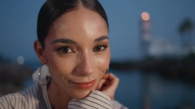 Muhteşem bir kadın, kameraya bakar ve doğaya nazikçe gülümser. Yaklaş. Akşam seti kenarında oturan kahverengi gözlü zarif bir kızın portresi. Mutlu şık esmer Alacakaranlıkta dinleniyor.