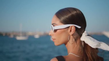 Güneşli okyanus kıyısında şık bir bayanın gözlüğünü çıkarışının portresi. Yaz plajında poz veren güzel genç bir kadın. Pahalı rıhtımda duran lüks model..