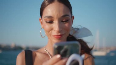 Güneşli rıhtımda cep telefonuyla selfie çeken bir portre kadın. Çekici genç bayan hafta sonu ayakta yüz resmi çekiyor. Sahilde cep telefonu kamerasına gülümseyen mutlu bayan turist.