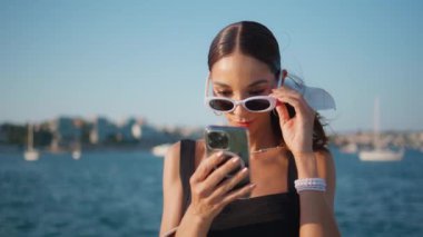 Güneşli toprak setinde cep telefonundan sosyal medyayı izleyen muhteşem bir kadın. Moda güneş gözlüklü mutlu, rahat kadın gülümseyerek akıllı telefon ekranına bakıyor. Zarif kız sahilde mesaj okuyor..