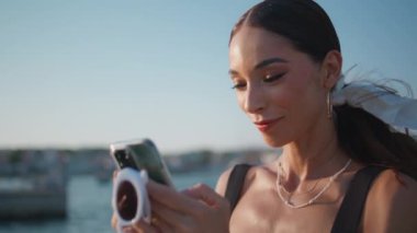 Yaz mevsiminde limanı kapatırken mesaj yazan rahat bir kız. Güneşli toprak setinde akıllı telefondan mesaj atan güzel, bronzlaşmış bir kadın. Moda bayan turist gezme mobil uygulaması deniz kıyısında dinleniyor.