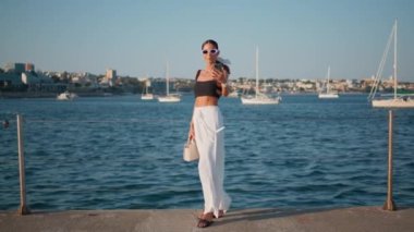 Yaz mevsiminde akıllı telefondan güzel bir turist kaydı videosu. Şatafatlı esmer kız dalgalı deniz suyunda cep telefonu kamerasıyla ayakta duruyor. Güneşli rıhtımda harika bir kadın blog yazıyor..