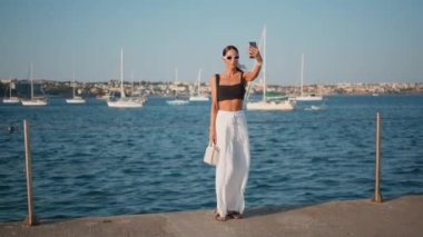 Deniz rıhtımında cep telefonu tutan gülümseyen kadın. Muhteşem stil sahibi kız lüks yatları olan güzel bir deniz manzarası kaydediyor. Güneş gözlüklü güzel bayan deniz manzaralı içeriği çekiyor..