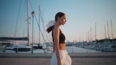 Beyaz lüks yatların yanında günbatımı seti gezen çekici bir model. Şık, kendine güvenen bir kadın, güneşli bir rıhtımda son moda kıyafetleriyle yürüyor. Muhteşem kız asfalt sahil yoluna çıkıyor yaz akşamı.