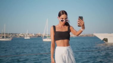 Bayan turist, Sunny Rıhtımı 'nda akıllı telefondan selfie çekiyor. Güneş gözlüklü çekici kadın rıhtımda duran cep telefonu kamerasına bakıyor. Havalı kız güzel deniz manzarasının önünde fotoğraf çekiyor..